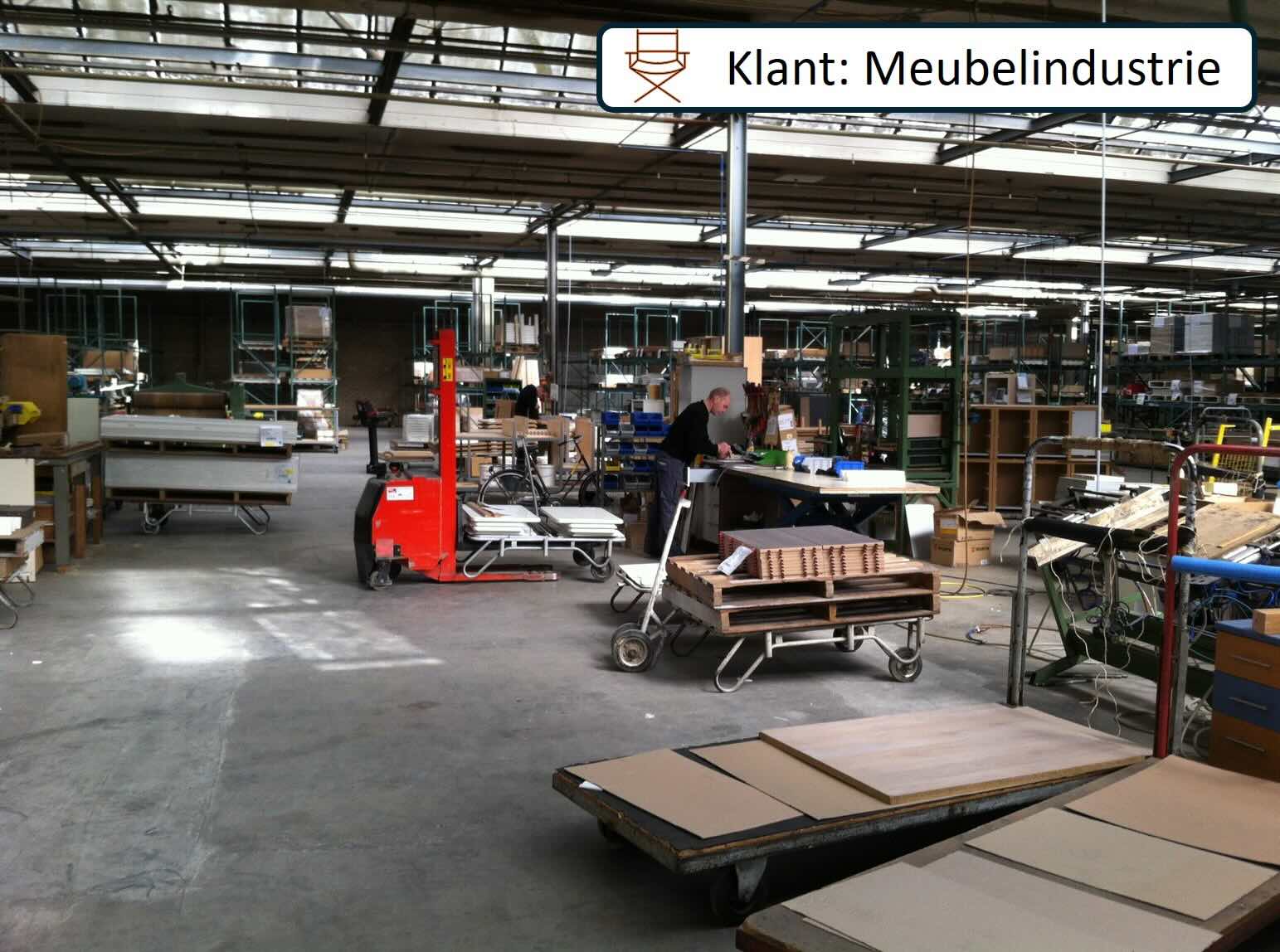 Medewerker werkt aan een tafel in een meubelproductiehal met houten onderdelen en transportkarren.