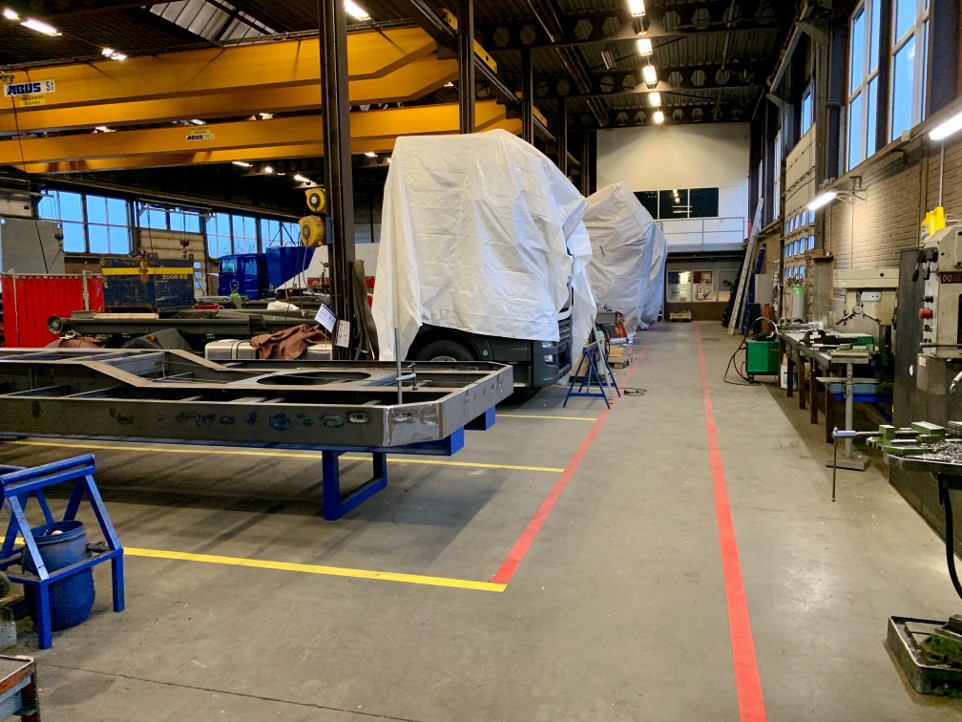 Productiehal met werkbanken, metalen onderdelen en afgedekte voertuigen.