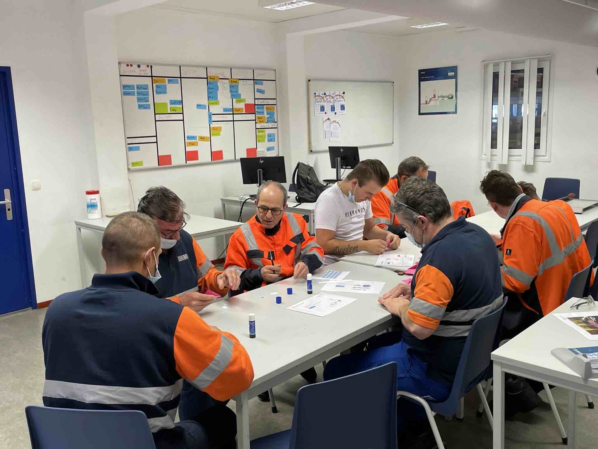 Medewerkers in werkkleding werken samen aan trainingsmateriaal aan een tafel.