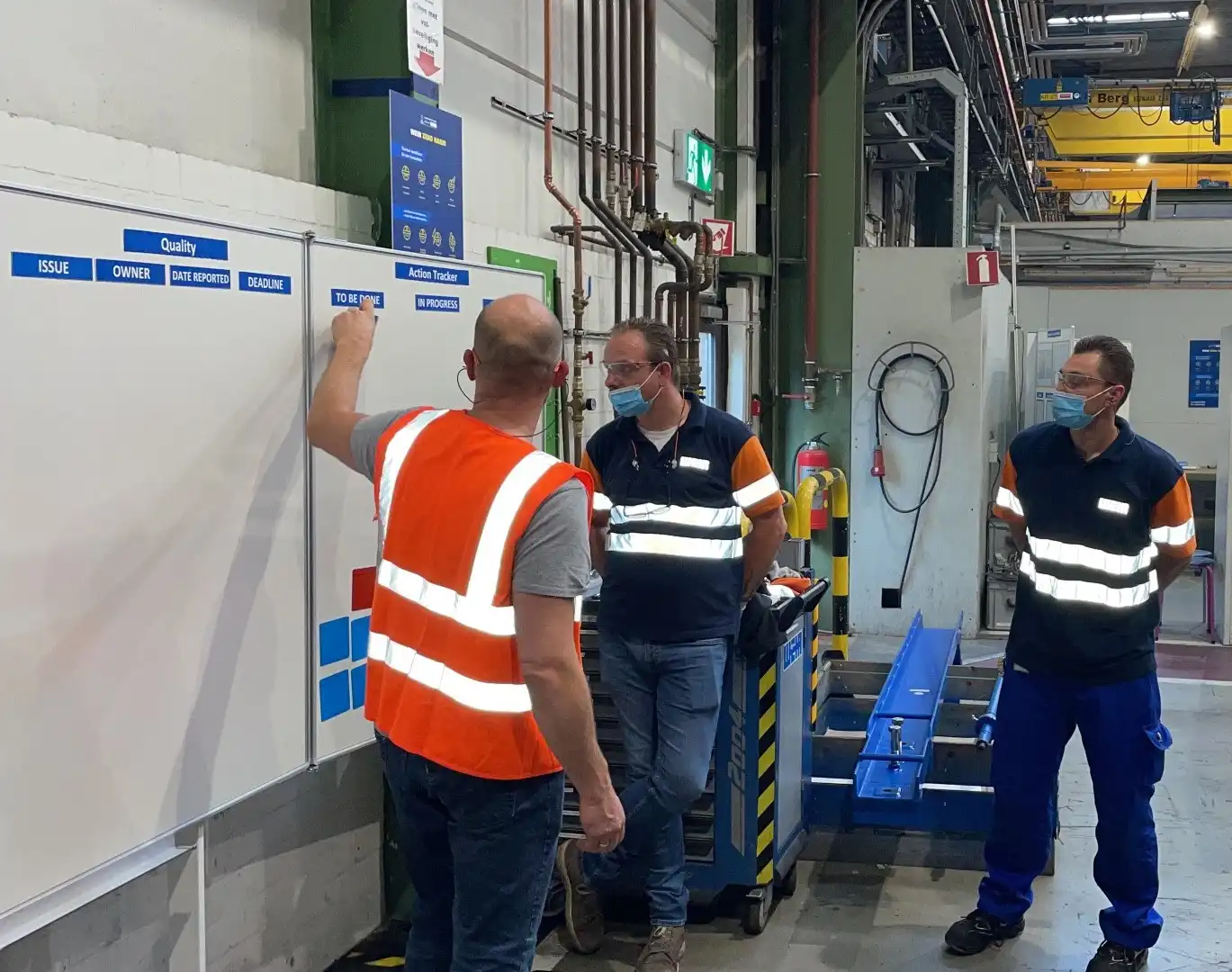Drie medewerkers bespreken acties bij een verbeterbord in de productiehal.